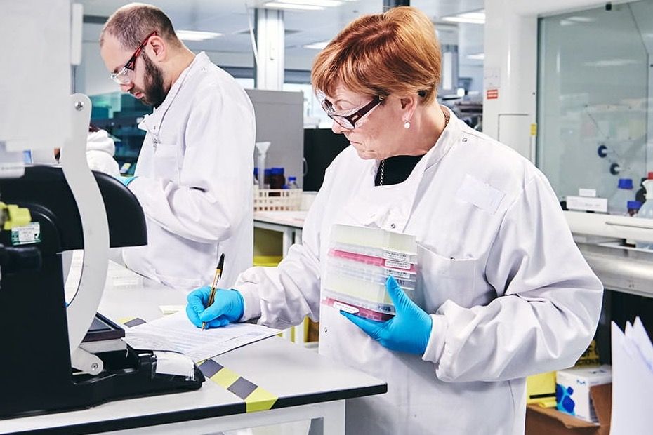 Due persone in camice rivedono un documento al banco di un laboratorio, una con un rack di campioni in mano